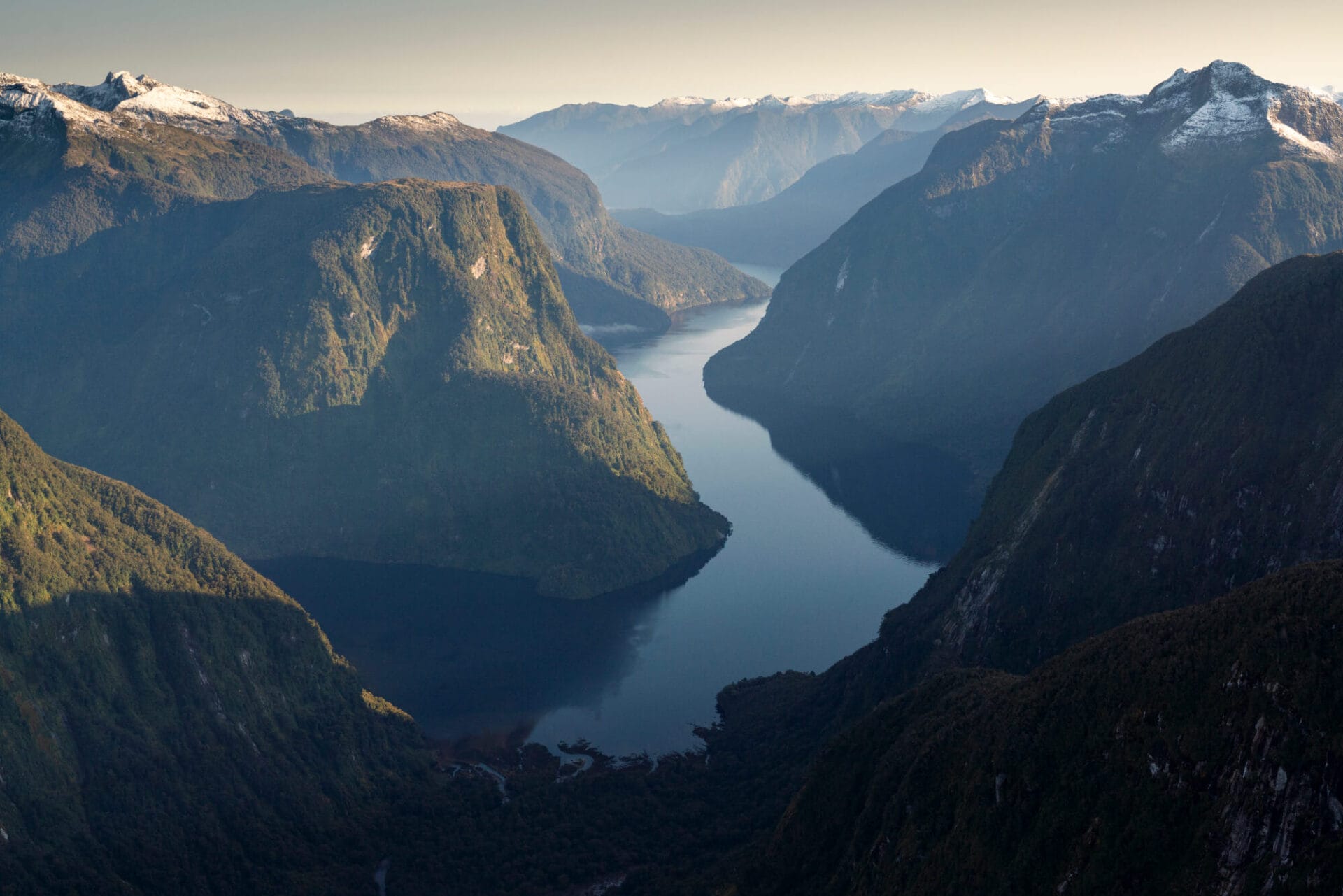 Aerial view of a serene fjord surrounded by rugged mountains with snow-capped peaks under a clear sky.