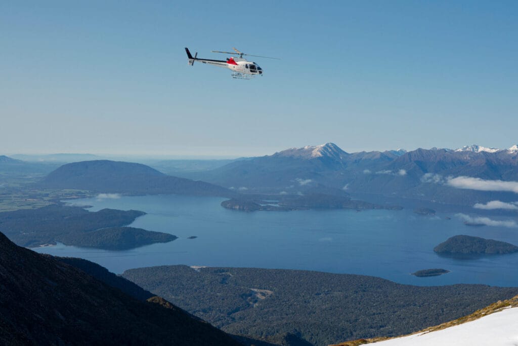 A helicopter flying over a scenic mountainous landscape with a large lake and several islands below, under a clear blue sky.