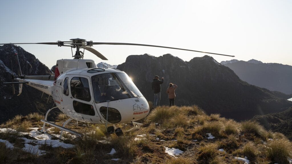 Two people standing near a helicopter on a mountain at sunset, with rugged peaks in the background.