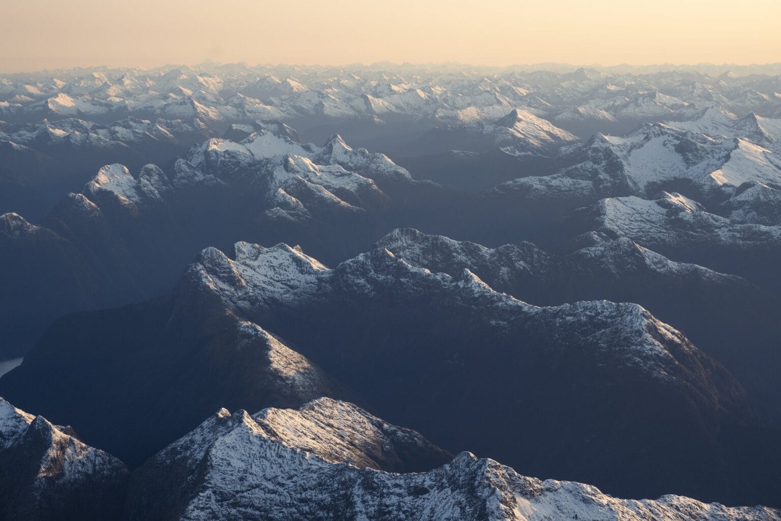 Aerial view of a mountain range at sunset, with peaks dusted in snow and valleys cast in shadows, bathed in soft golden light typical of a Fiordland experience.