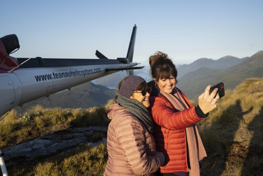 Two women taking a selfie next to a Fiordland helicopter on a mountain at sunset.