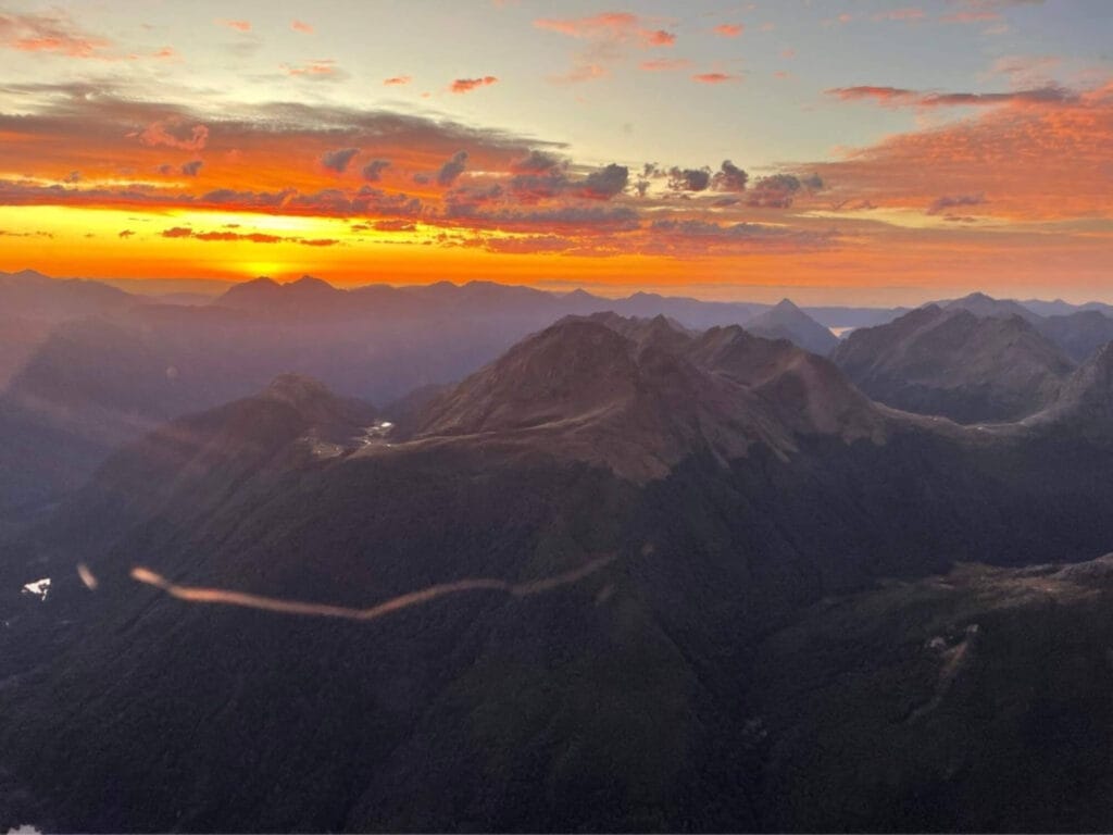 Aerial view of a mountain range during sunrise, showing vibrant orange and yellow skies with shadowed peaks and valleys.