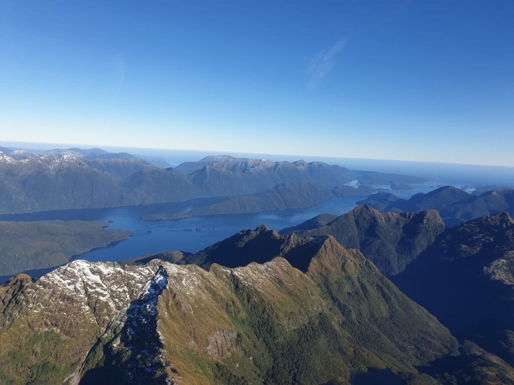 Aerial view of Dusky Sound. A rugged mountain range with snow patches, overlooking a serene blue lake and distant mountains under a clear sky.