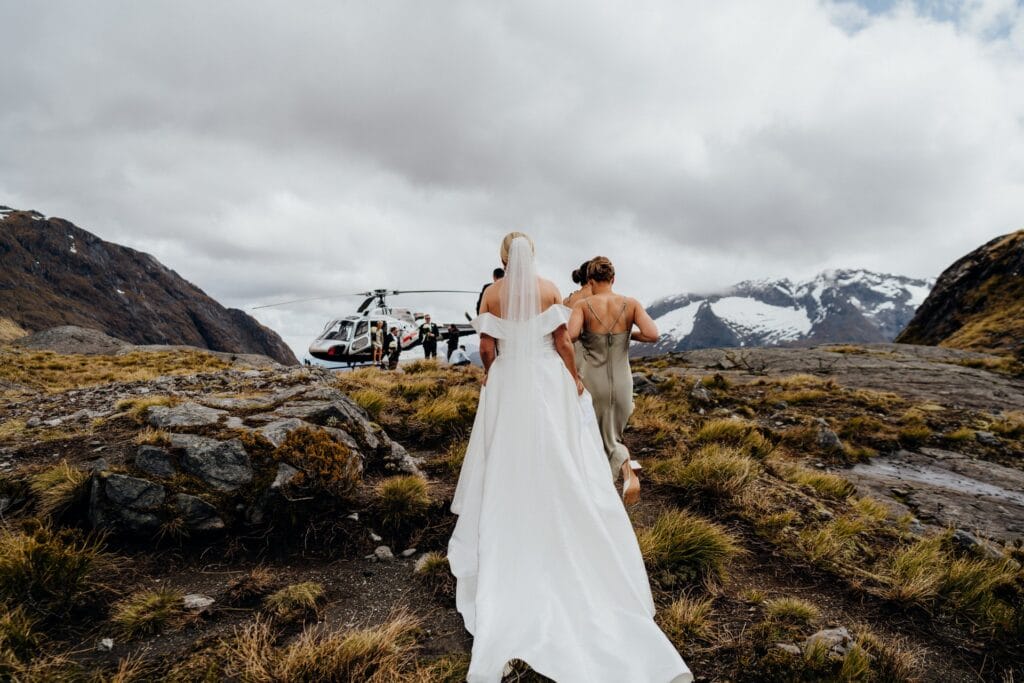 A bride and a bridesmaid walking towards a helicopter on a rugged mountain landscape, with snowy peaks in the background.
