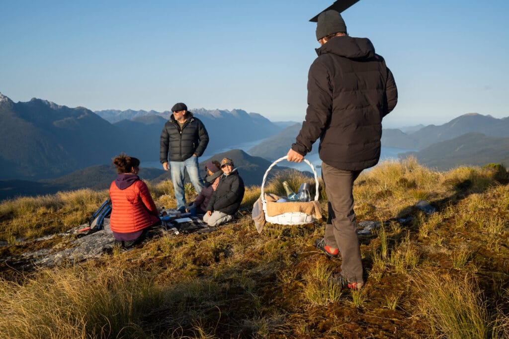 Group of people enjoying a picnic on a mountain at sunset, with expansive views of distant mountain ranges.