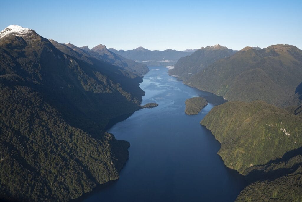 Aerial view of a serene fjord surrounded by forested mountains with snowy peaks under a clear blue sky.