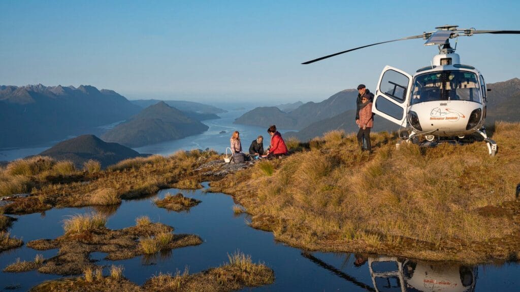 Three people enjoy a picnic next to a helicopter on a mountain overlooking a chain of lakes and distant mountains in Fiordland at sunset.