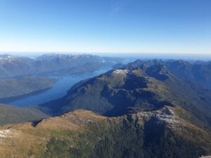 Aerial view of a fiordland landscape with clear blue waters and rugged terrain under a bright blue sky.