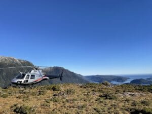 Helicopter landing on a grassy mountain ridge in Fiordland with a panoramic view of a deep valley and distant mountains under a clear blue sky.