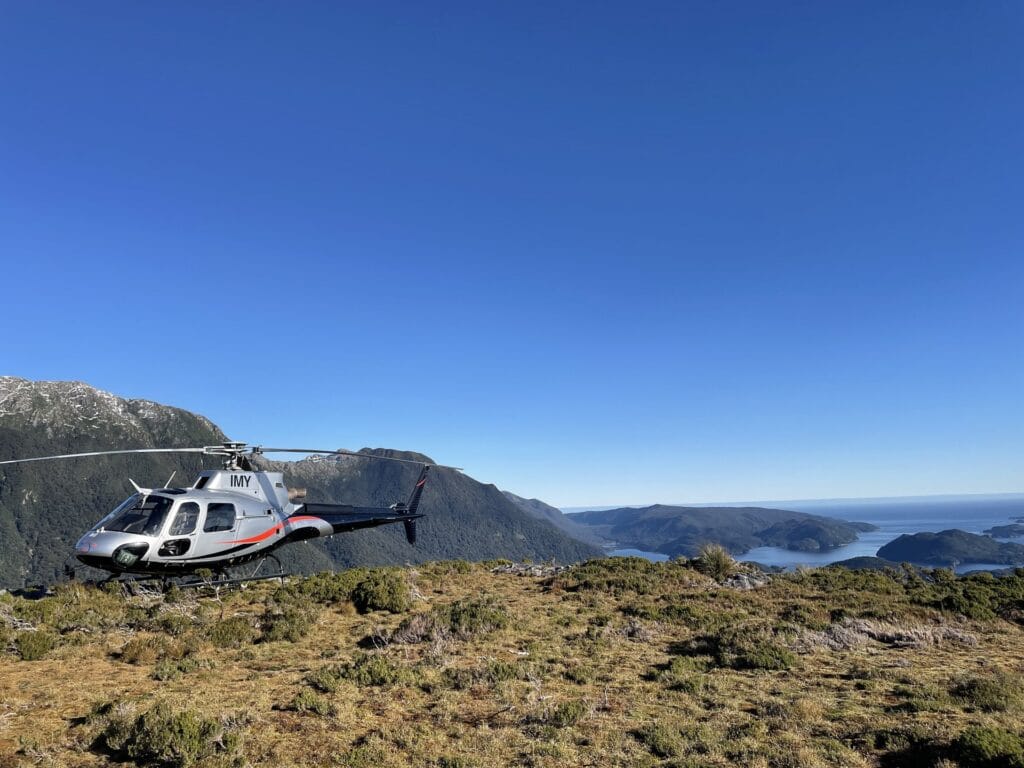 Helicopter landed on a mountain overlooking a fjord under clear blue skies.