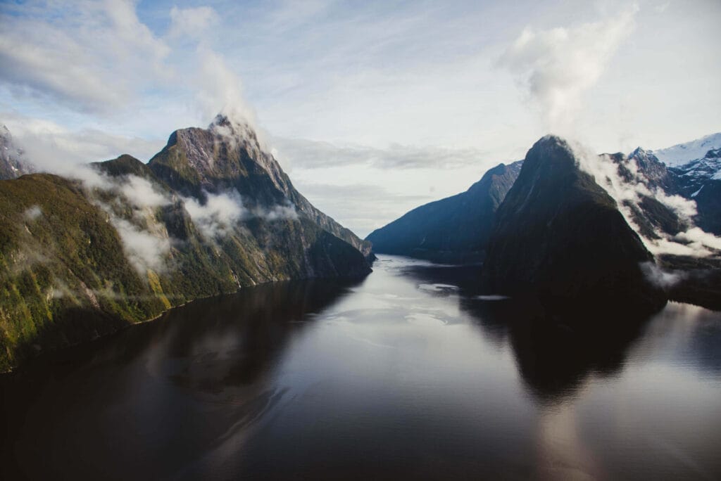 A scenic view of Milford Sound in New Zealand, showcasing towering mountains reflected in tranquil waters under a partly cloudy sky.