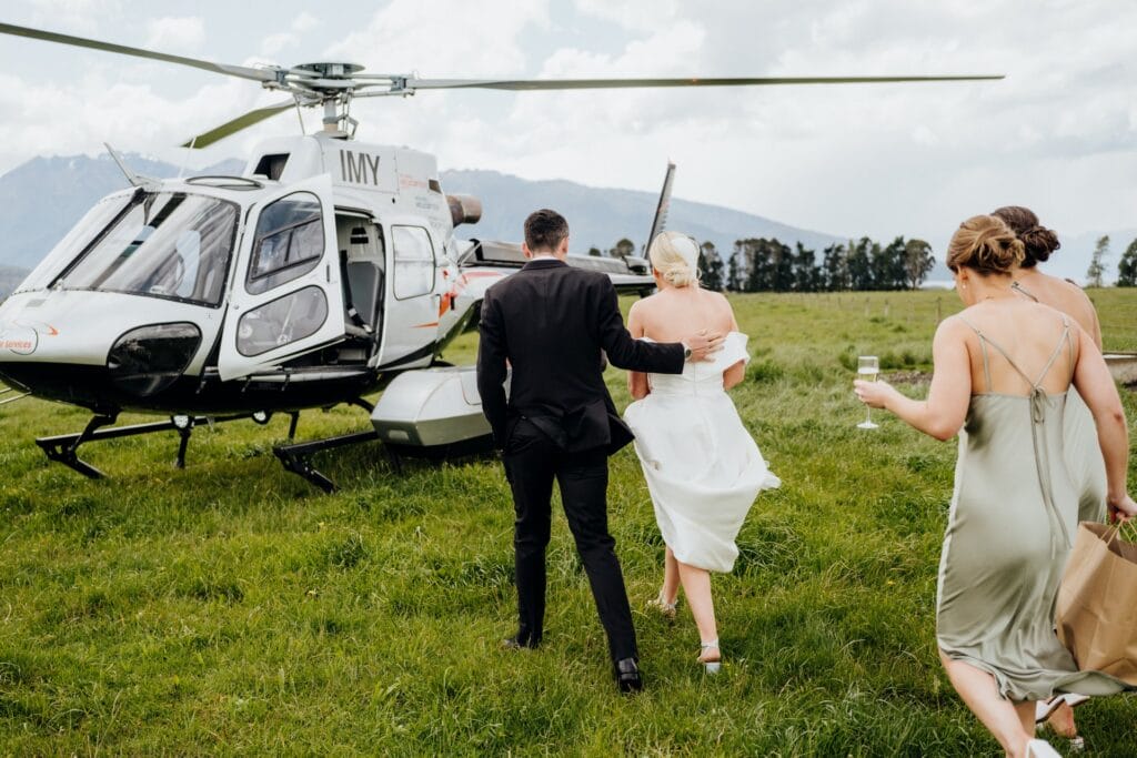 A bride and groom, accompanied by a guest, walking towards a helicopter in a grassy field, indicating a luxurious wedding departure.