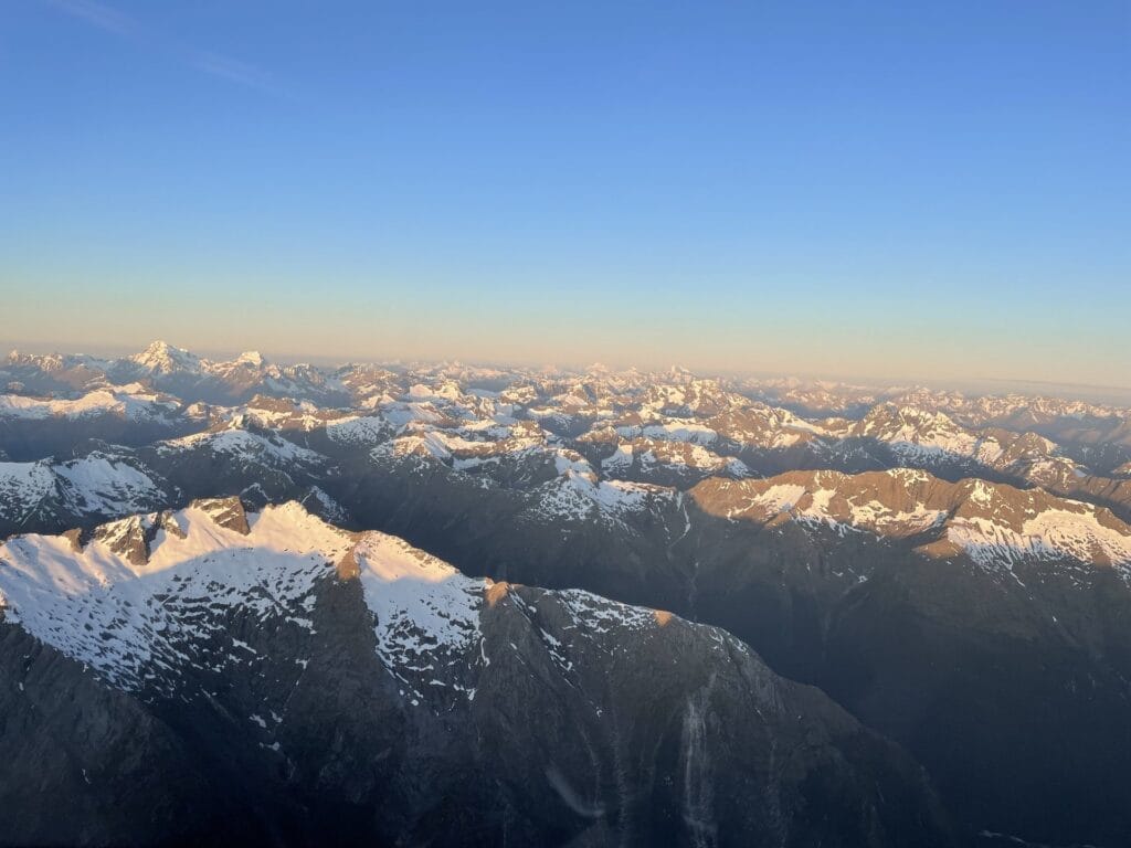 Aerial view of snow-capped mountain peaks bathed in golden sunlight during a clear sunrise, captured from a Fiordland helicopter.