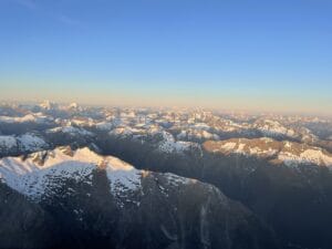 Aerial view of snow-capped mountain peaks bathed in golden sunlight during a clear sunrise, captured from a Fiordland helicopter.