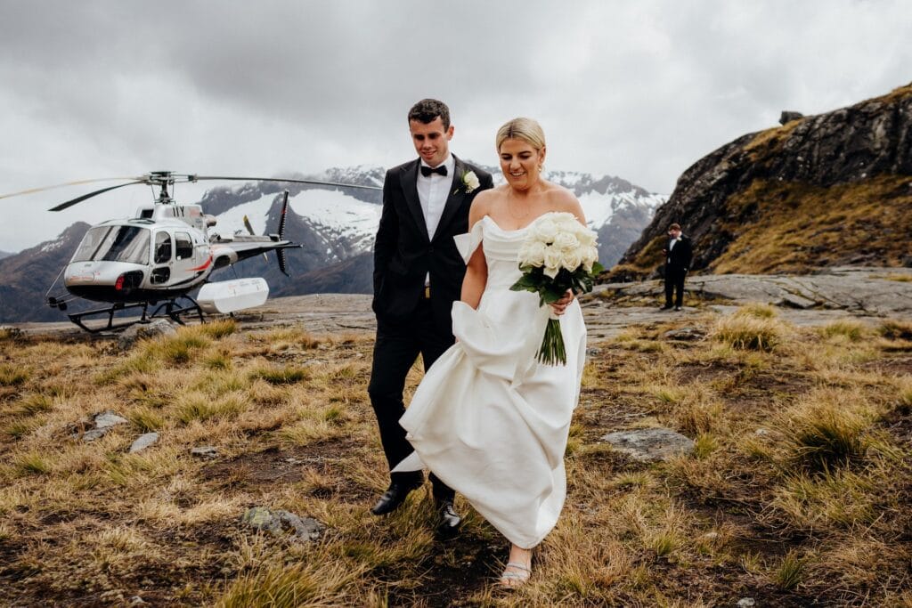 A newly married couple, the bride in a white gown and the groom in a black tuxedo, walk hand in hand near a helicopter on a mountainous landscape with sparse vegetation and snow-capped peaks in the background.