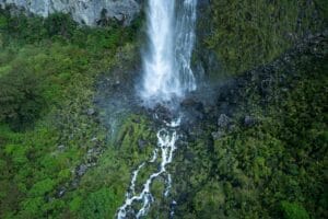 Aerial view of a tall waterfall in Fiordland cascading down a rocky cliff into smaller streams surrounded by lush green vegetation.