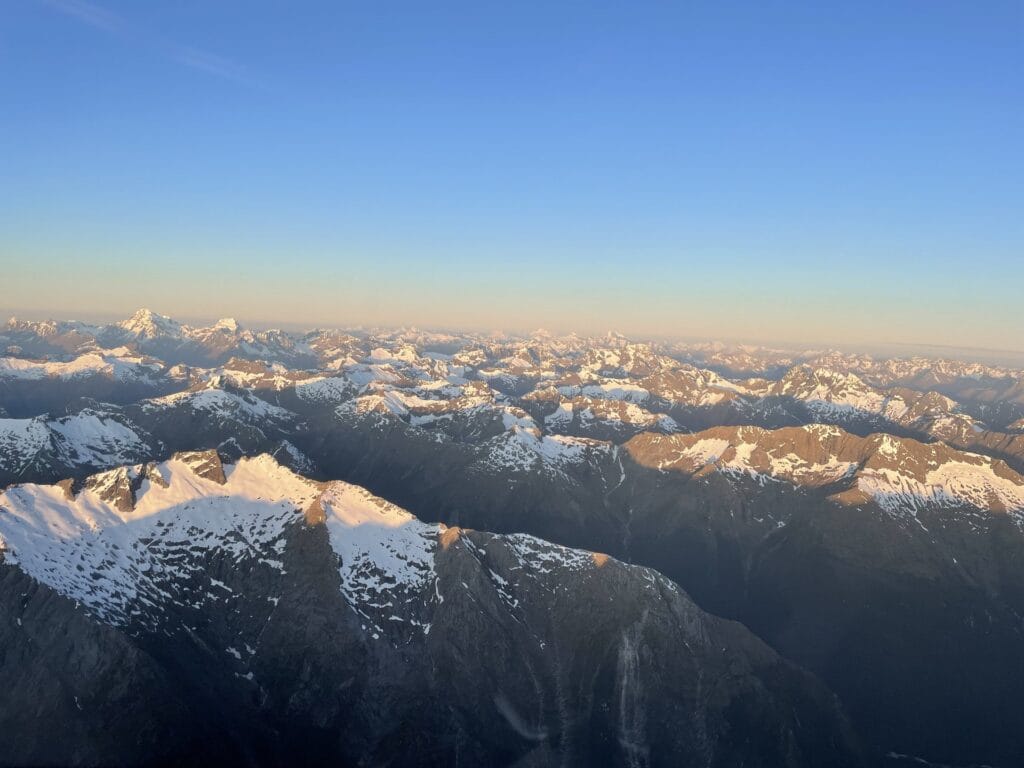 Sunrise over a rugged mountain range, casting a golden light on snow-capped peaks against a clear blue sky.