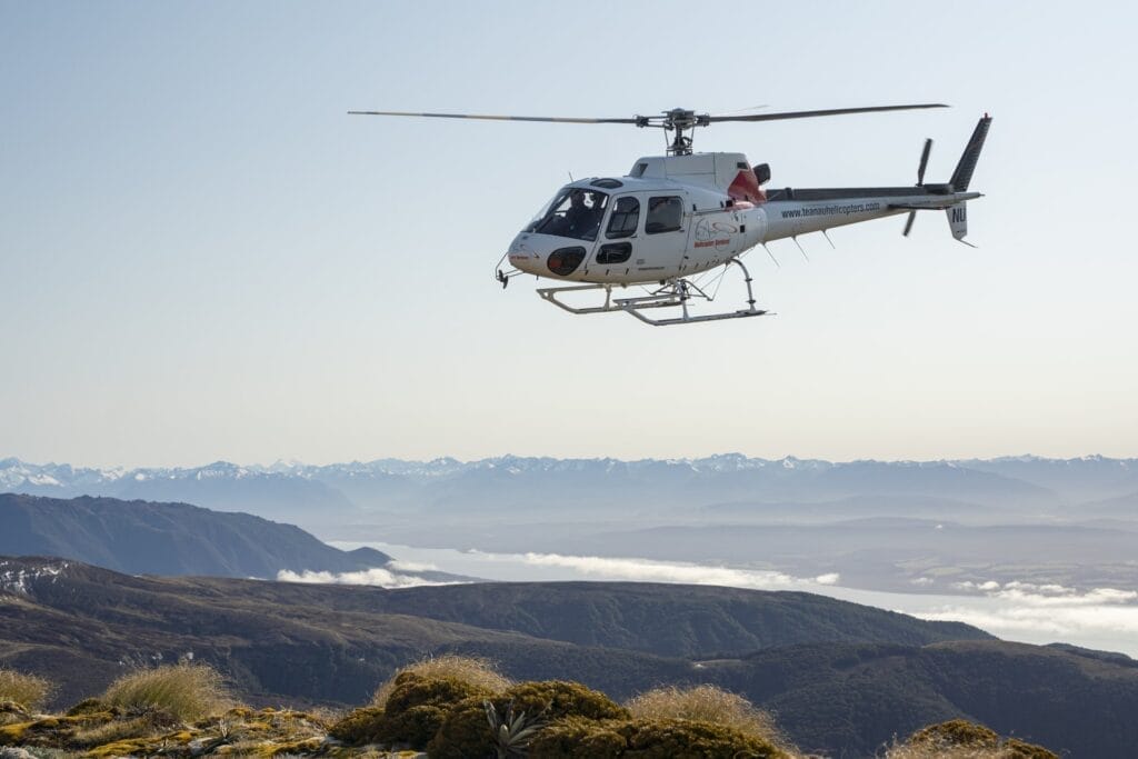 A helicopter flies over a mountainous landscape with visible distant snow-capped peaks and a valley shrouded in clouds during a Kepler heli hike.