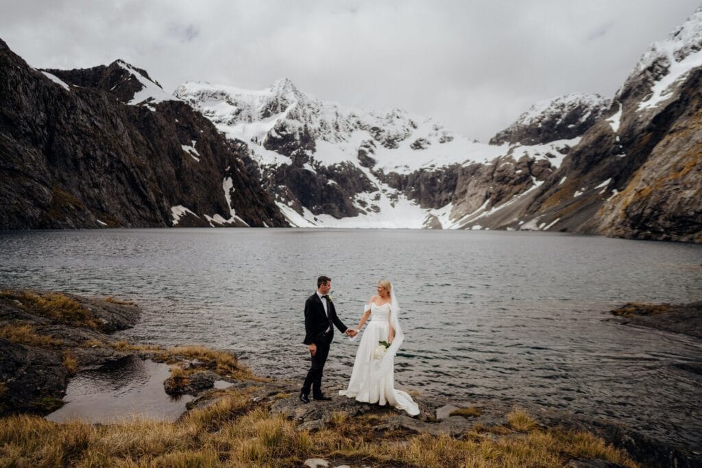 A bride and groom holding hands near a mountain lake with snowy peaks in the background.