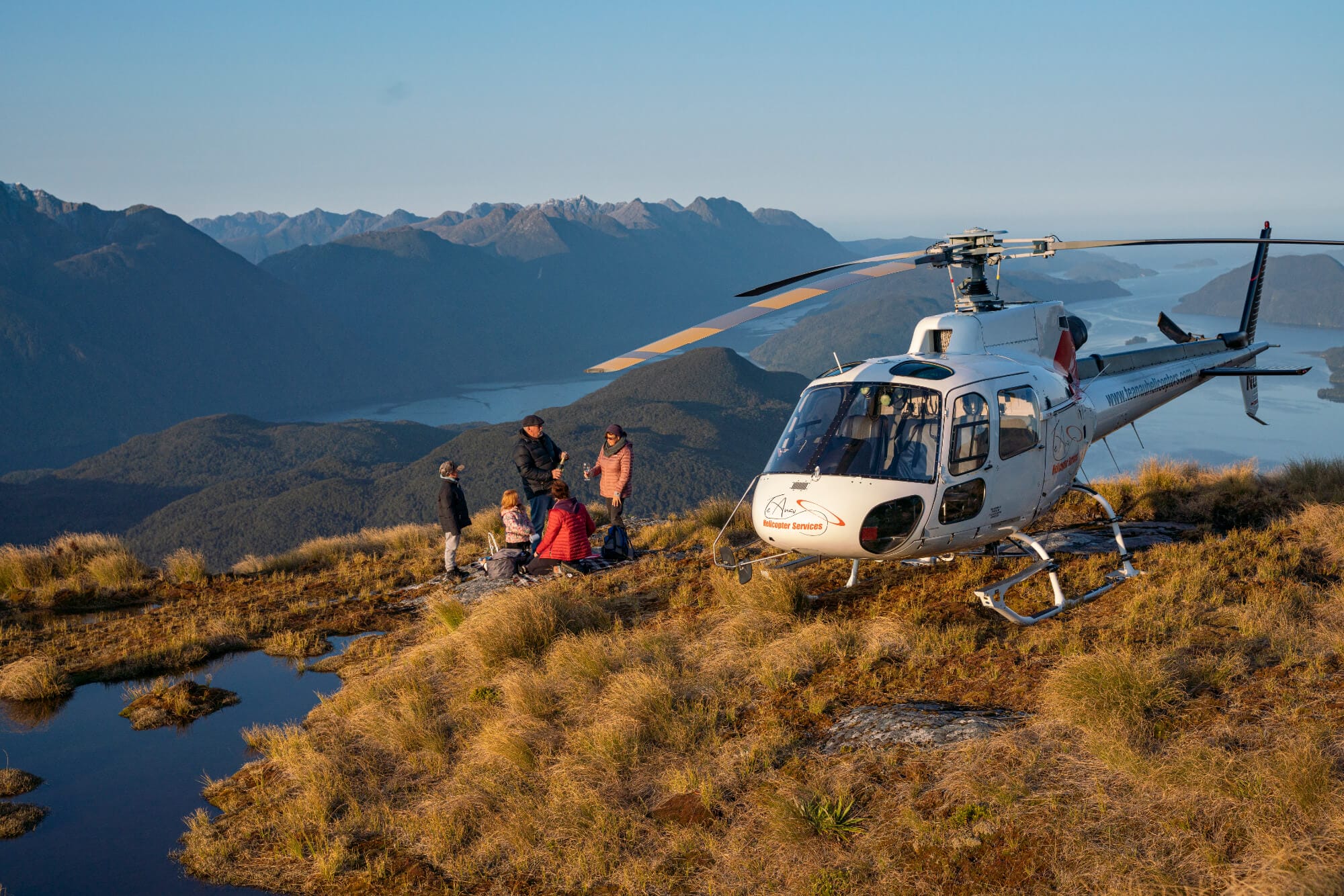 A group of people sitting near a helicopter on a mountain summit, with panoramic views of distant mountains and a lake.