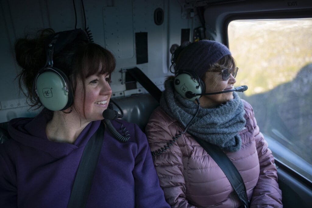 Two women wearing headsets while seated inside a Fiordland helicopter, looking out the window, dressed in warm clothing.