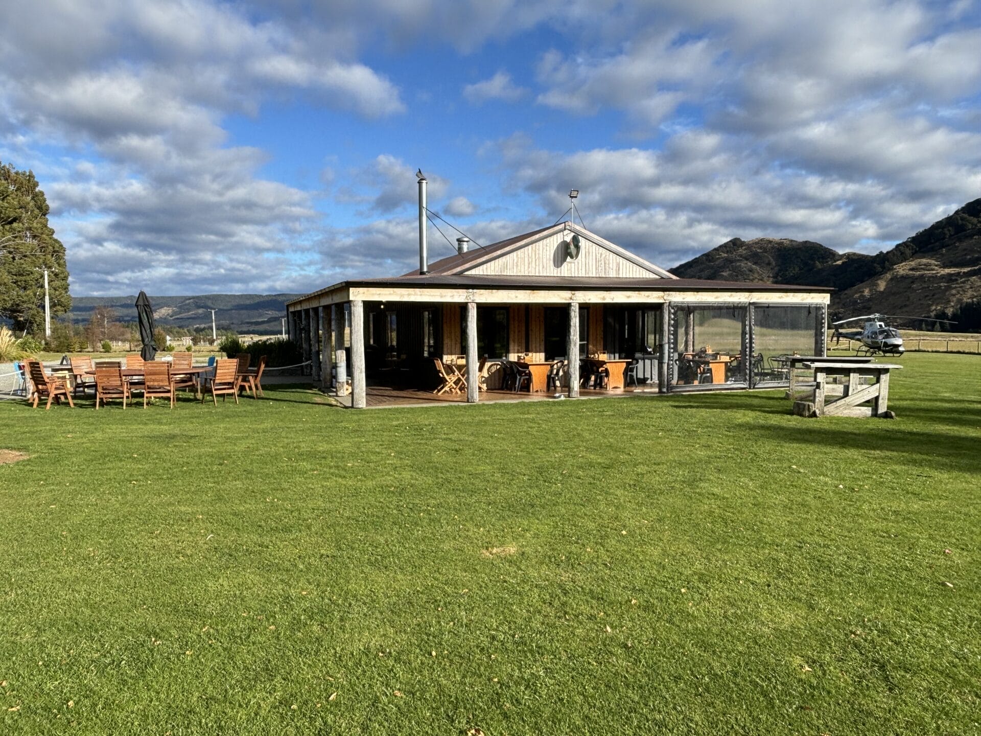 A rustic wooden building with large windows stands on a grassy lawn with neighboring mountains and cloudy skies in the background, capturing the essence of a Fiordland trip. An outdoor dining area with wooden tables and chairs is visible to the left.
