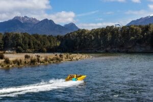 Fiordland Jet cruises on a river with forested banks and mountains in the background under a partly cloudy sky.