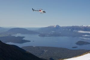 A helicopter flies over a mountainous landscape with a large lake and scattered islands below, capturing the breathtaking views of a Fiordland trip. The sky is clear, and the serene scene unfolds from an elevated vantage point.