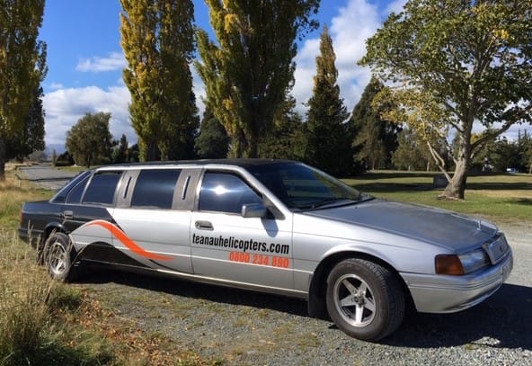 A silver stretch limousine parked in a field with tall trees in the background and advertising decals for a helicopter service on its side.