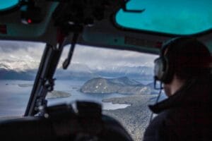 From the helicopter cockpit, a passenger wearing headphones gazes at the breathtaking mountains and shimmering waters below, experiencing the unparalleled beauty offered by Manapouri Tours.