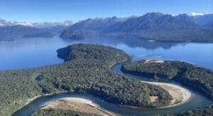 Experience the breathtaking aerial view of a lush, forested peninsula embraced by the expansive Lake Manapouri. With distant mountains under a pristine blue sky, this picturesque scene is perfect for those seeking unparalleled beauty on their Manapouri flight.