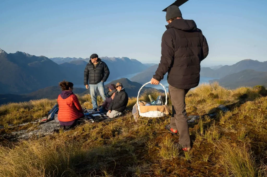 Four people in outdoor gear are having a picnic on a grassy mountain slope, with one person approaching carrying a basket. The mountainous landscape extends into the background under a clear sky.