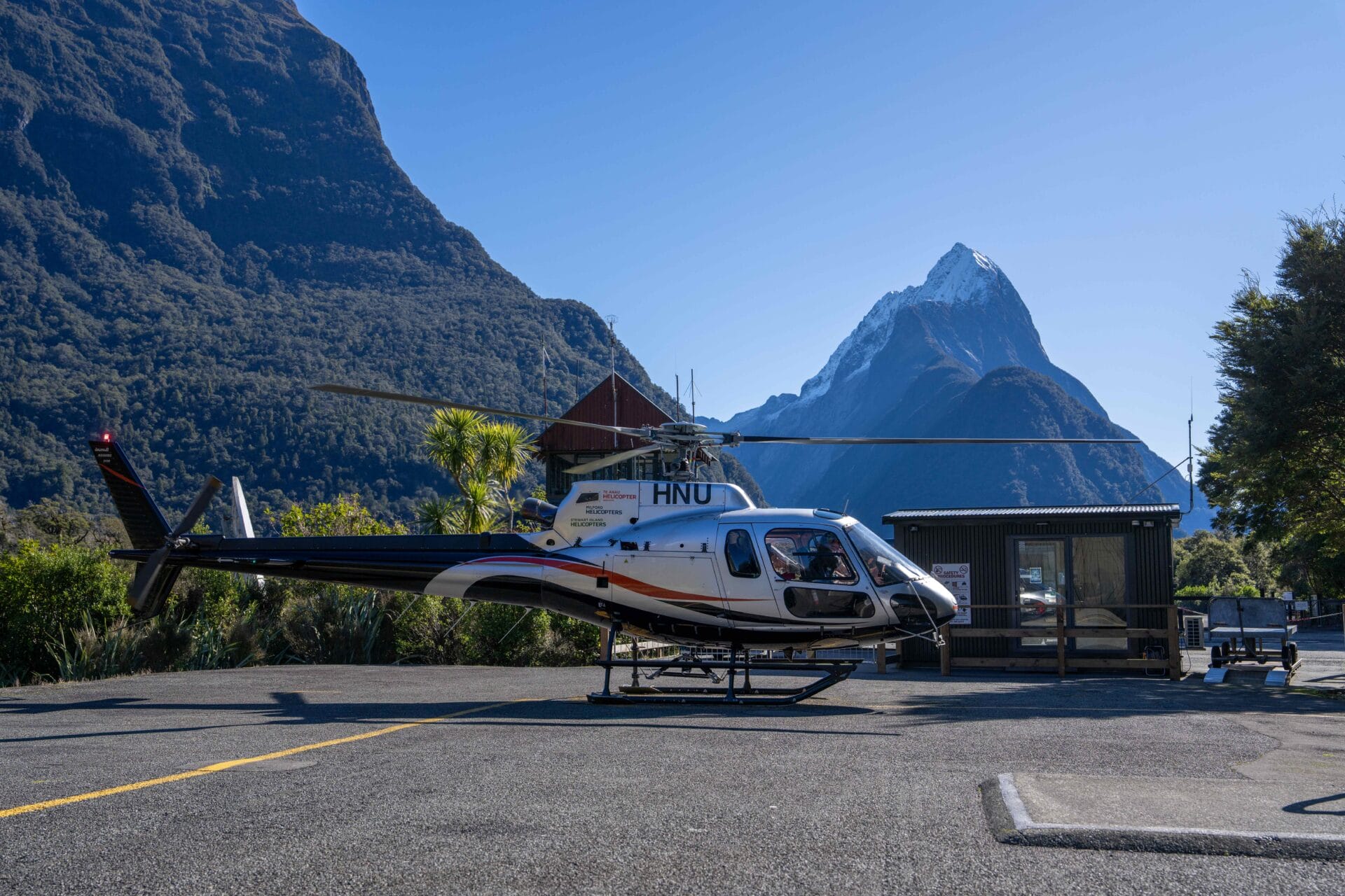 A helicopter is parked on a helipad near a small building, with a tall mountain peak and tree-covered slopes in the background under a clear sky.